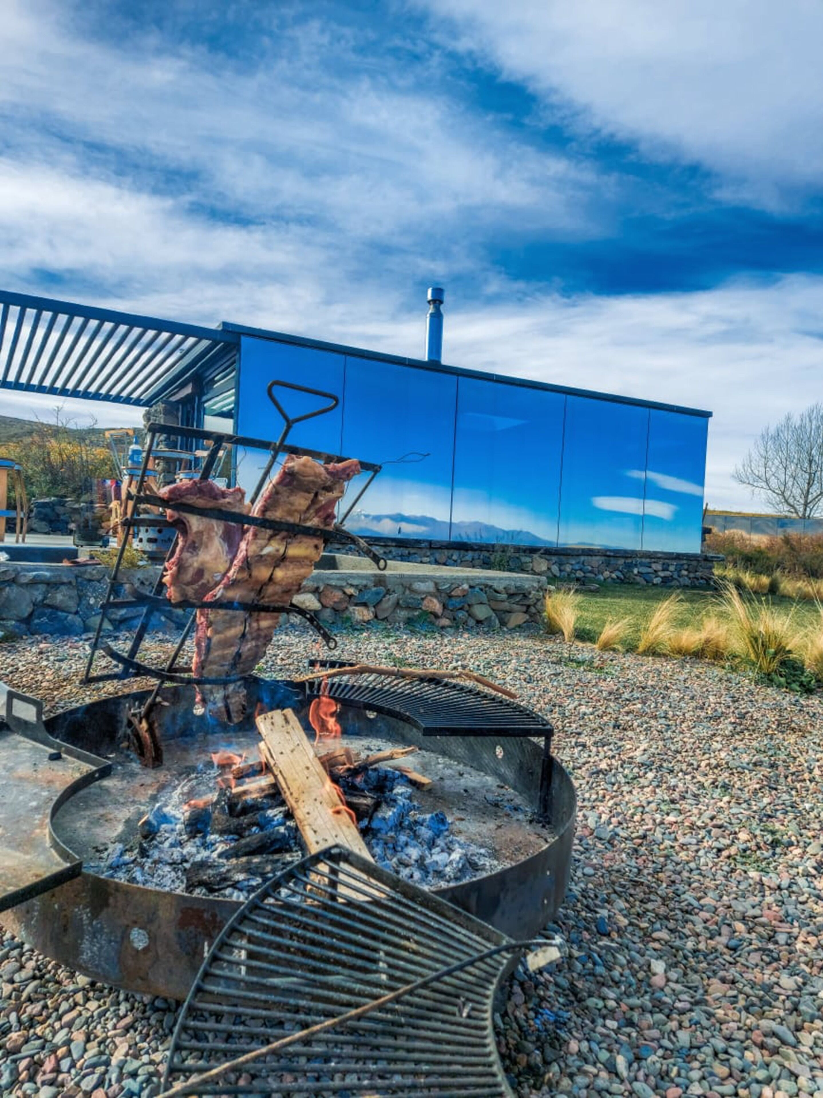 Una parrilla al aire libre con carne, un muro de piedra y un edificio azul al fondo.