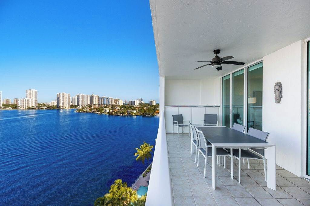 A balcony with a dining table and chairs, overlooking a body of water with buildings in the distance.