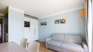 A modern living room with a sofa, a dining table, and chairs. There is a kitchen area with cabinets and a refrigerator. - Némo - Appartement avec Terrasse (Courseulles-sur-Mer)