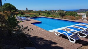 Et rektangulært swimmingpool med blåt vand, omgivet af en tr æterrasse. Hvide liggestole med blå hynder står på terrassen. I baggrunden ses en klar blå himmel og et fjerntliggende kystlandskab med bygninger og bakker.