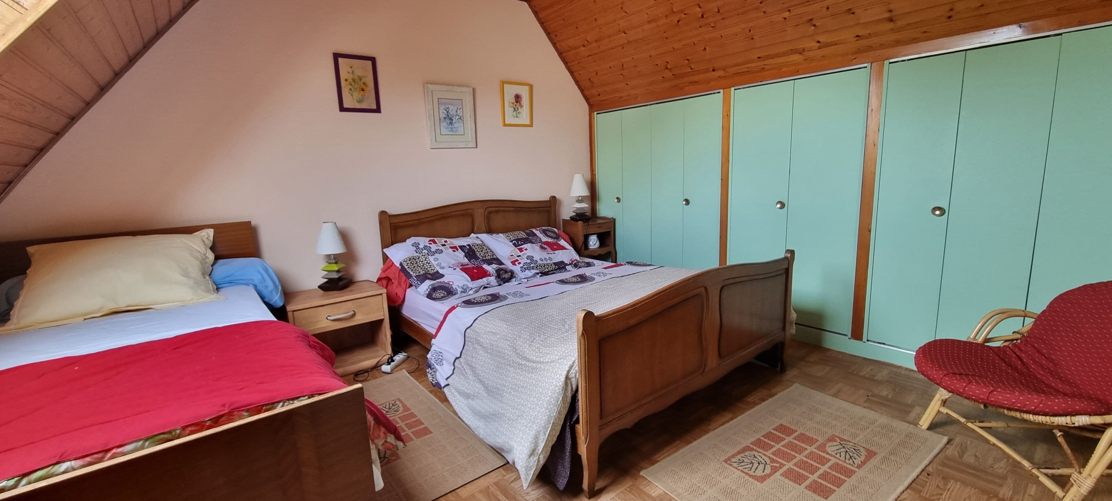 Een slaapkamer met een houten plafond, een bed met quilt, een nachtkastje met lamp, een garderobe en een rieten stoel.