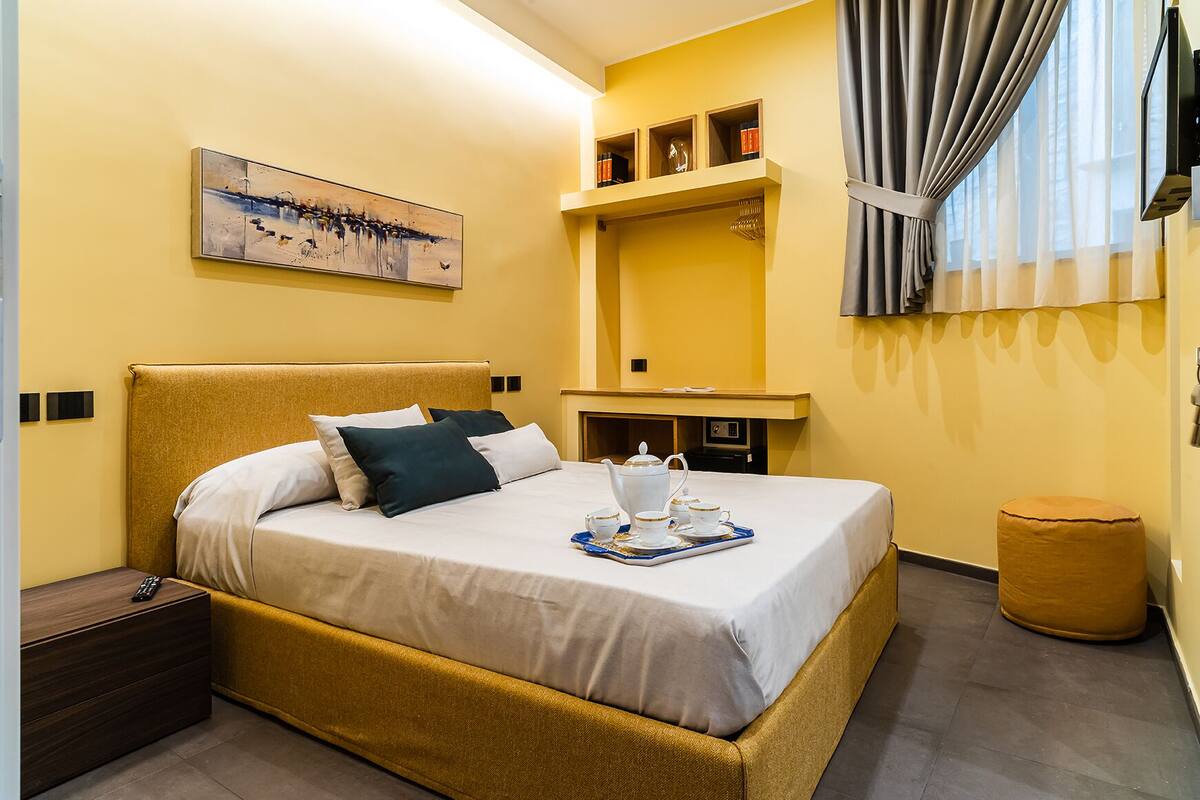 A hotel room with a bed, bedside table, a small table with a tray of tea, a painting on the wall, a shelf with books and decorative items, and a window with curtains.