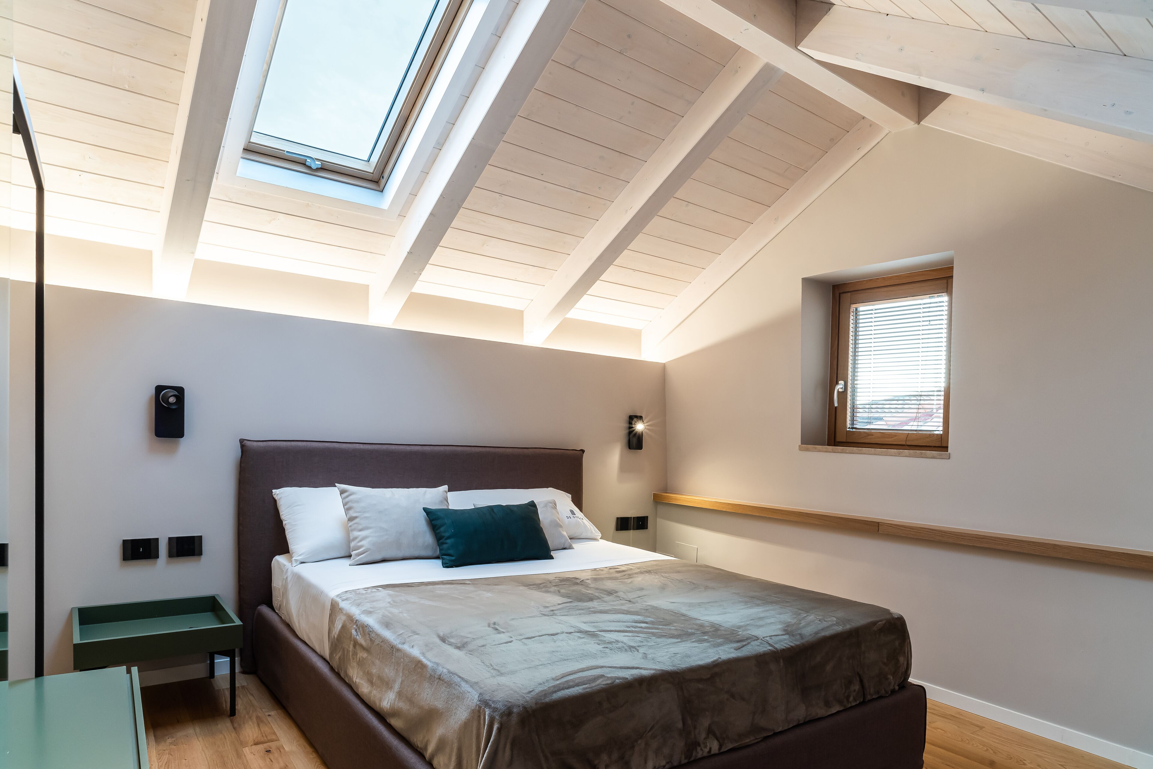 A bedroom with a skylight, a bed with pillows, a bedside table, and a window with blinds.