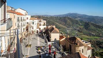 Una calle estrecha flanqueada por edificios, un área pavimentada con mesas al aire libre y una vista de un paisaje montañoso al fondo.
