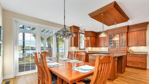 Une cuisine spacieuse avec des meubles en bois, un coin repas doté d’une grande table et de chaises, et une vue dégagée grâce aux portes vitrées.