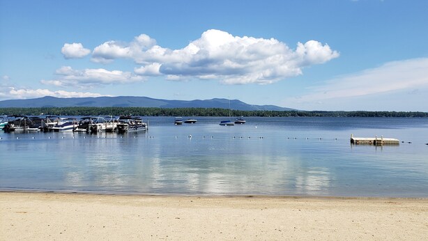 A calm lake with boats docked on the left, a sandy beach in the foreground, and a forested shoreline on the right.