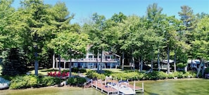 Location de vacances au bord d’un lac, avec un quai, un bateau et une grande maison entourée d’arbres.