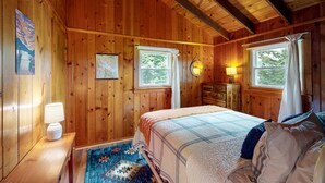 A wooden cabin bedroom with a bed, two windows, a nightstand with a lamp, and a dresser.