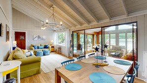 A spacious living room with a wooden dining table, blue cushions, and a large window offering a view of the outdoors.