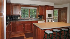 A kitchen with wooden cabinets, a large island with stools, a built-in oven, and a window overlooking a forest.