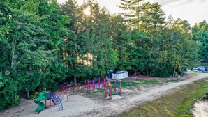 A playground with climbing structures and a slide, surrounded by dense trees and a dirt path. - Directly Across Street From Suissevale Winnipesaukee Beach (Moultonborough)