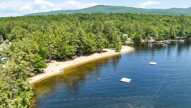 A lake surrounded by dense forest and a sandy beach with boats. - Directly Across Street From Suissevale Winnipesaukee Beach (Moultonborough)