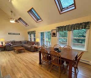 Uma sala de estar com sofá, mesa de centro, mesa de jantar com cadeiras, ventilador de teto, claraboias e uma placa que diz 'LAKE HOUSE'.