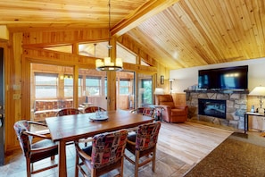 A spacious living area with a wooden ceiling, a stone fireplace, a television mounted on the wall, and a large window providing natural light.