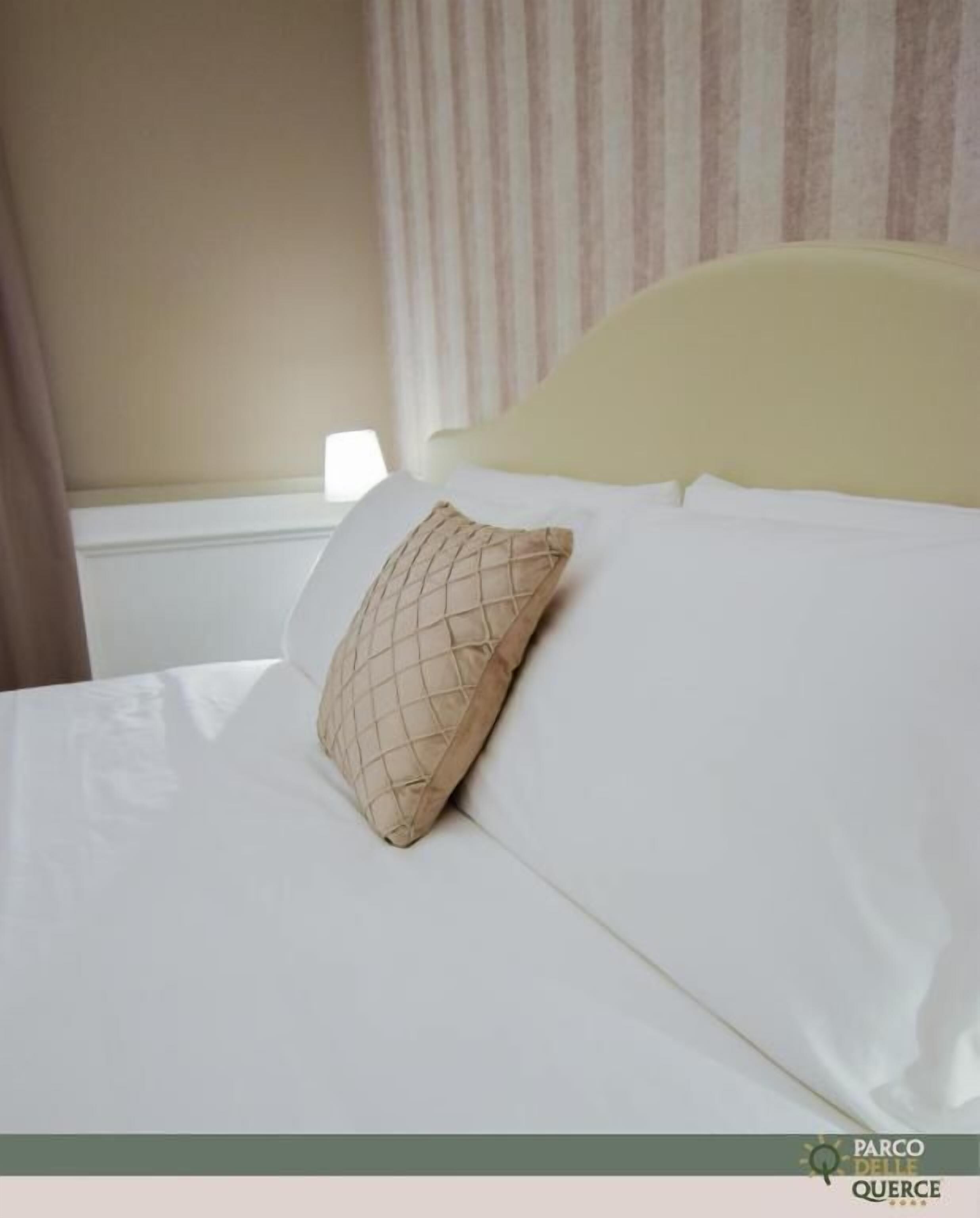 Una cama con ropa blanca y una almohada color beige, una ventana con cortinas y un cabecero de tono claro.