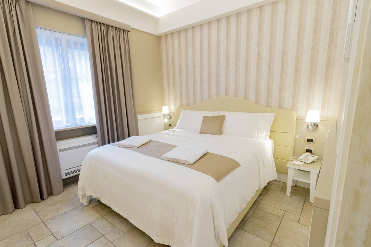 Habitación de hotel con una cama grande, una mesita de noche con lámpara, teléfono y una ventana con cortinas.
