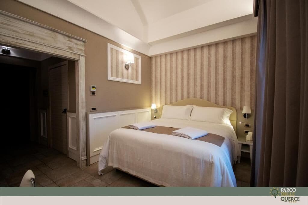 Quarto de hotel com uma cama grande, uma mesa de cabeceira, uma luminária fixada na parede e uma porta que dá para outro quarto.