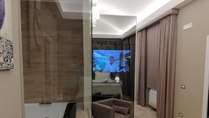 Moderni hotellihuone, jossa on suuri taulutelevisio, sohva ja sängyn vieressä yöpöytä.