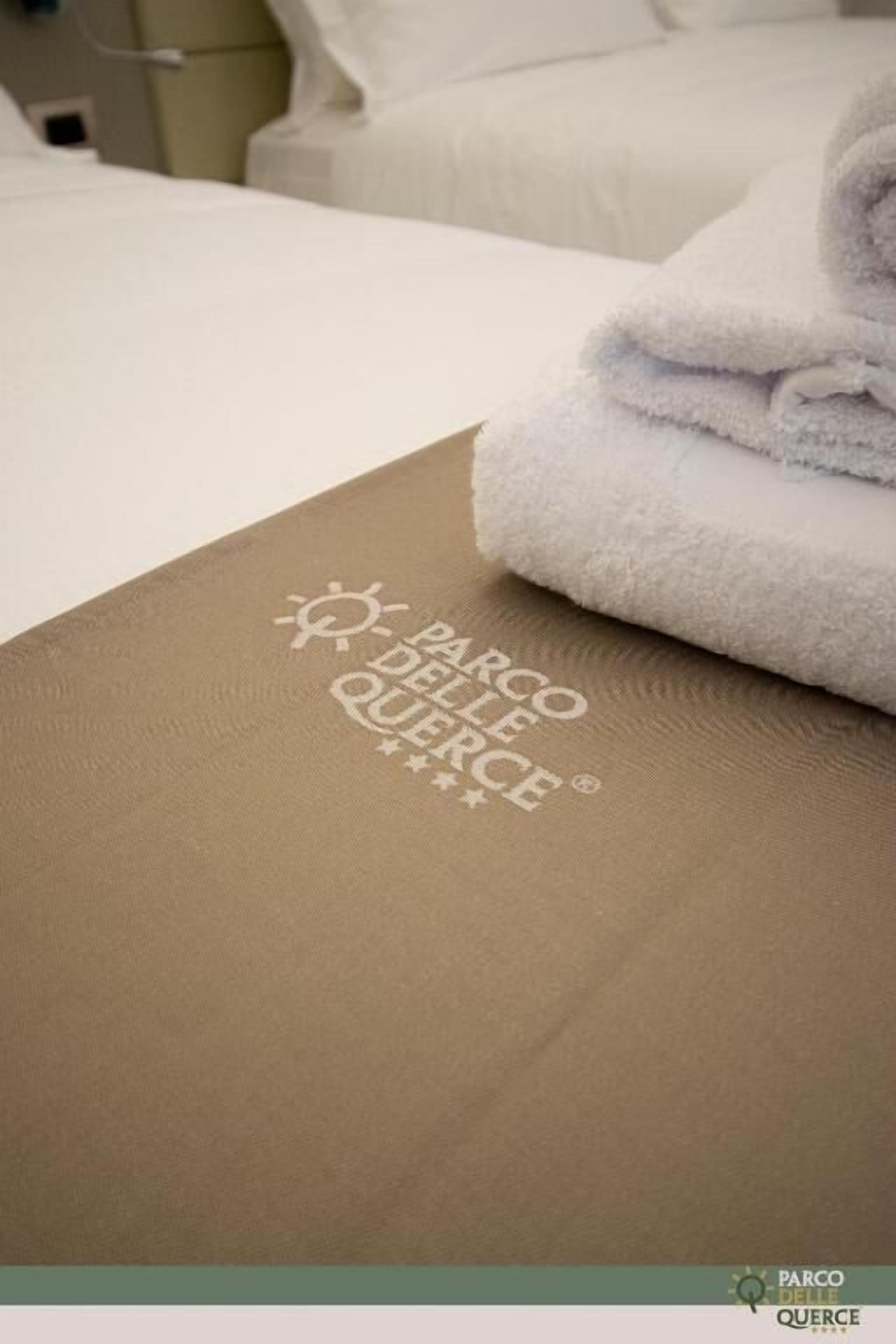 A neatly made bed with a folded white towel and a brown placemat with the text "PARCO DELLE QUERCE".