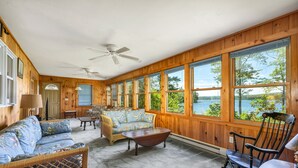 A cozy cabin interior with a large window overlooking a lake, a ceiling fan, and a wicker sofa. - Winnipesaukee Home PLUS Apartment For 10 Beach Dock Sunsets AUG 29TH SPECIAL (Moultonborough)