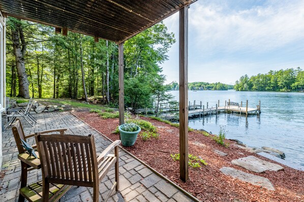 A covered deck with wooden chairs, a stone pathway, and a view of a lake with a dock.