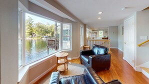 A spacious living room with a large window overlooking a body of water, a wooden floor, and a black leather sofa. - Winnipesaukee Waterfront in Meredith for 10 (Meredith)