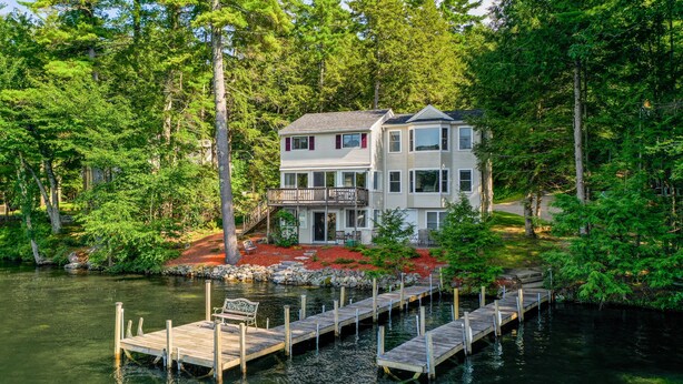 A two-story house with a dock extending into a lake, surrounded by dense trees. - Winnipesaukee Waterfront in Meredith for 10 (Meredith)