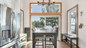 A modern kitchen with a stainless steel refrigerator, a wooden dining table with chairs, and large windows offering a view of trees and a lake.