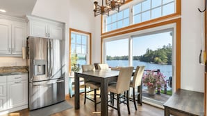 A kitchen with a large stainless steel refrigerator, a dining table with chairs, and a view of a lake through large windows.