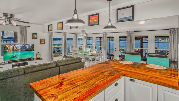 A spacious living area with a wooden bar, large windows offering ocean views, a ceiling fan, and a television displaying a scenic landscape. - READY TO RELAX?  Lake front house w/ beautiful Sunsets on Lake Greenwood (cross Hill)