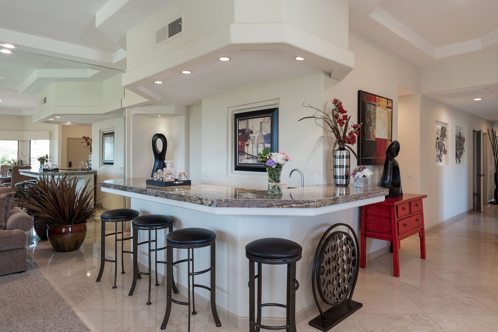 A modern kitchen with a central island, bar stools, a red side table, and decorative items.