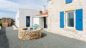 Un bâtiment en pierre avec des volets bleus, un petit espace extérieur aménagé avec des sièges et une fontaine en pierre.