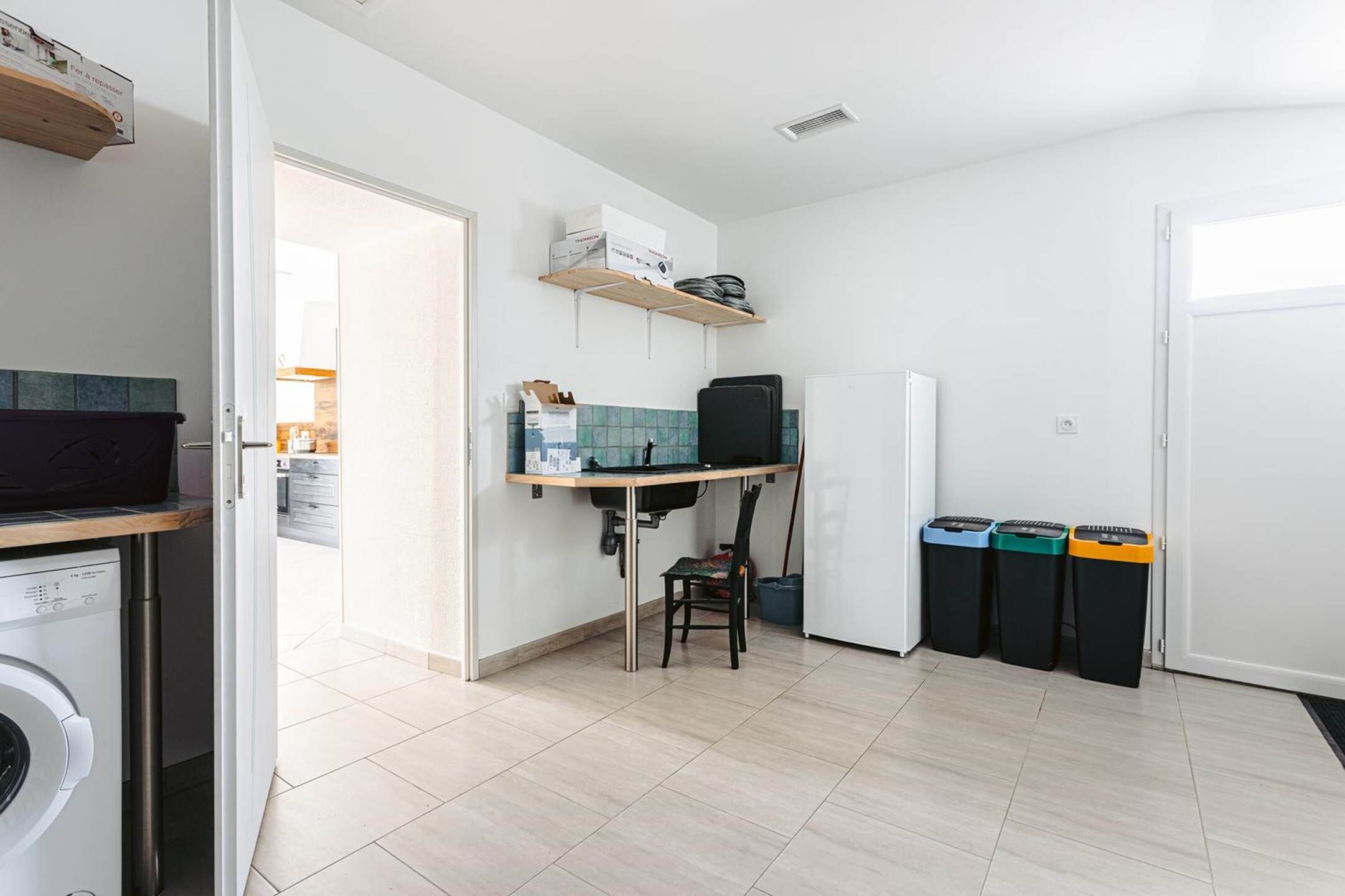 A compact kitchen with a washing machine, a small dining table, and a chair. There are three trash bins and a refrigerator.