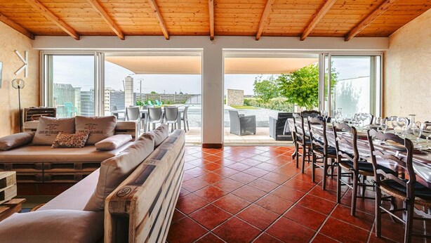 Une pièce spacieuse avec un plafond haut, de grandes fenêtres, un coin repas avec des chaises et des tables, et un coin salon avec des canapés.