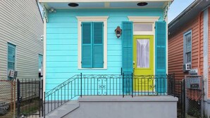 Una casa de dos pisos con fachada azul turquesa brillante, una puerta amarilla y un pequeño porche con barandilla de hierro forjado negro.