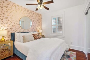 Une chambre à coucher comprenant un lit, un ventilateur de plafond, une fenêtre avec des stores et une table de chevet avec une lampe.
