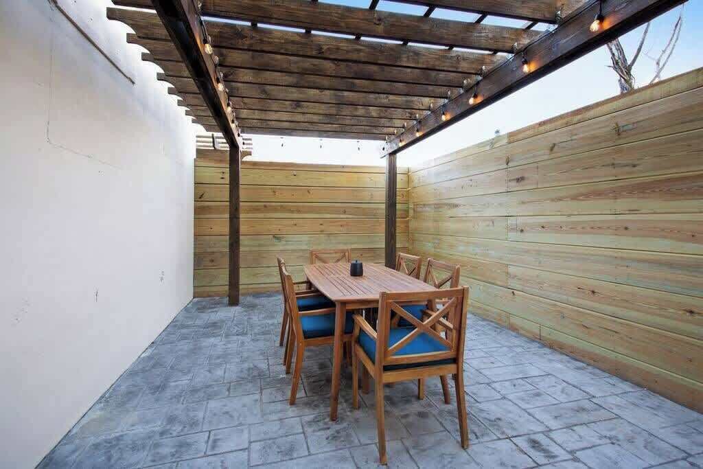 A covered patio area with a wooden table and chairs, a pergola overhead, and a wooden wall.
