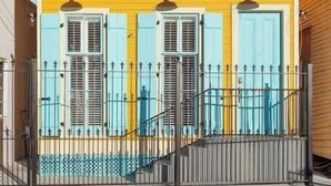 Un edificio de dos pisos con paredes amarillas y contraventanas azules, que presenta una entrada con reja y una pequeña escalera que lleva al piso superior.
