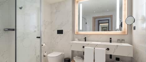 A modern bathroom with a glass shower enclosure, a marble-tiled floor, a large mirror with a wooden frame, a white toilet, and a vanity with a wooden shelf holding towels.