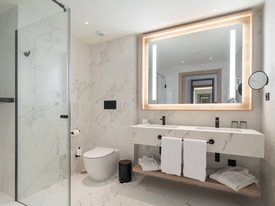 A modern bathroom with a glass shower enclosure, a marble-tiled floor, a large mirror with a wooden frame, a white toilet, and a vanity with a wooden shelf holding towels.