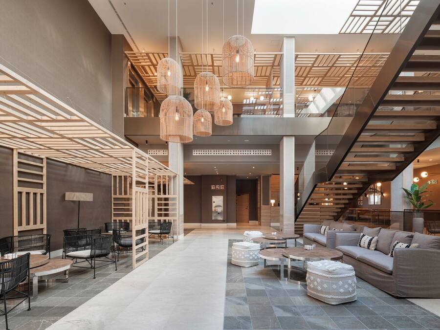 Un vestíbulo amplio con diseño moderno, que cuenta con una zona de estar central con sofás y mesas, una escalera con barandilla de madera y lámparas colgantes.