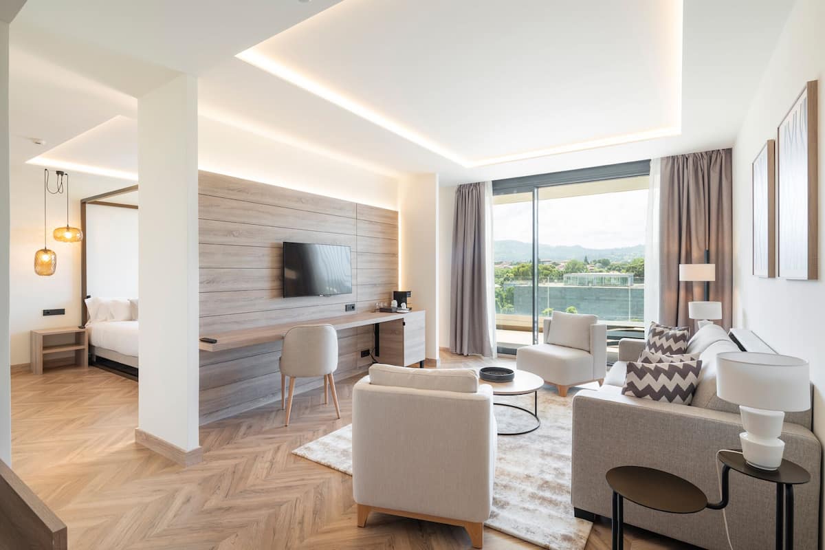 Quarto de hotel moderno com uma grande janela que oferece vista para um corpo de água e colinas. Há uma TV de tela plana fixada em uma parede de madeira, uma área de estar com sofá e cadeiras, e uma pequena mesa com abajur.