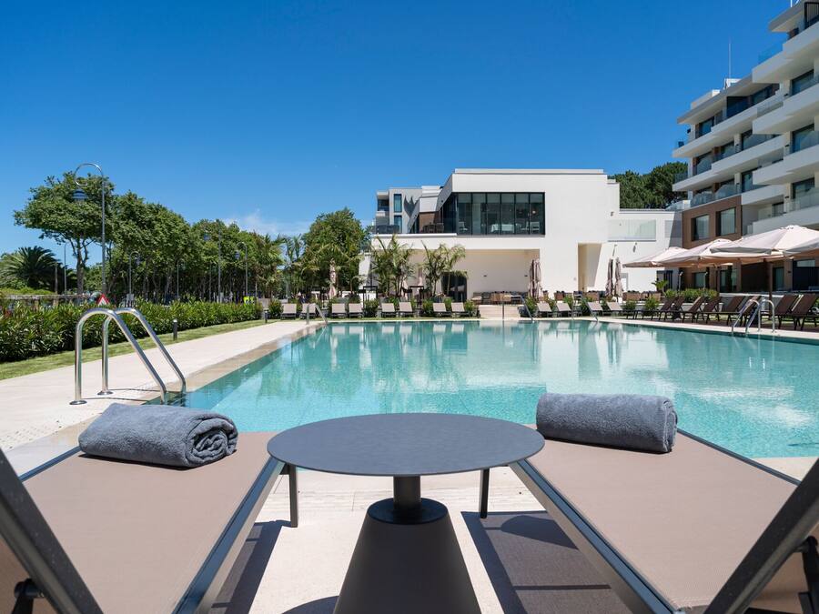 A modern hotel pool area with lounge chairs and a table, surrounded by greenery and a clear blue sky.