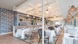 Un restaurante con mesas blancas y sillas de mimbre, pared de ladrillo y lámparas colgantes.