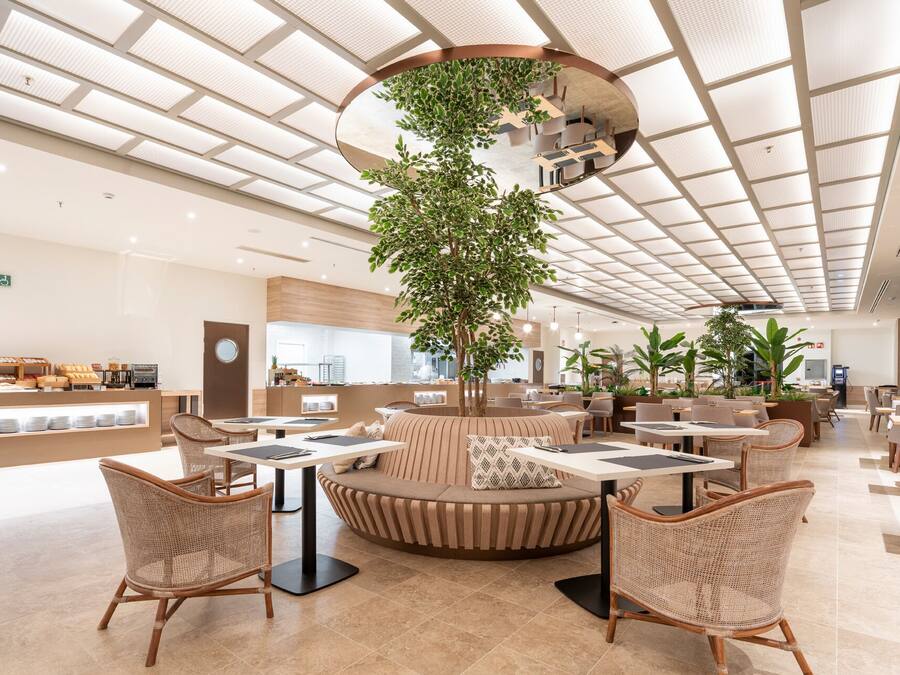 A spacious indoor area with round tables and wicker chairs, a central seating area with a tree, and a ceiling with a grid pattern.