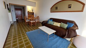 Ein Wohnzimmer mit einem braunen Sofa, einem weißen Couchtisch und einem blauen Teppich. Im Hintergrund befindet sich ein Esstisch mit Stühlen.
