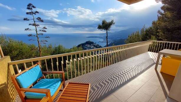 Ein Balkon mit einem Holztisch und blauen Sitzgelegenheiten, der einen schönen Blick auf das Meer und ferne Berge bietet.