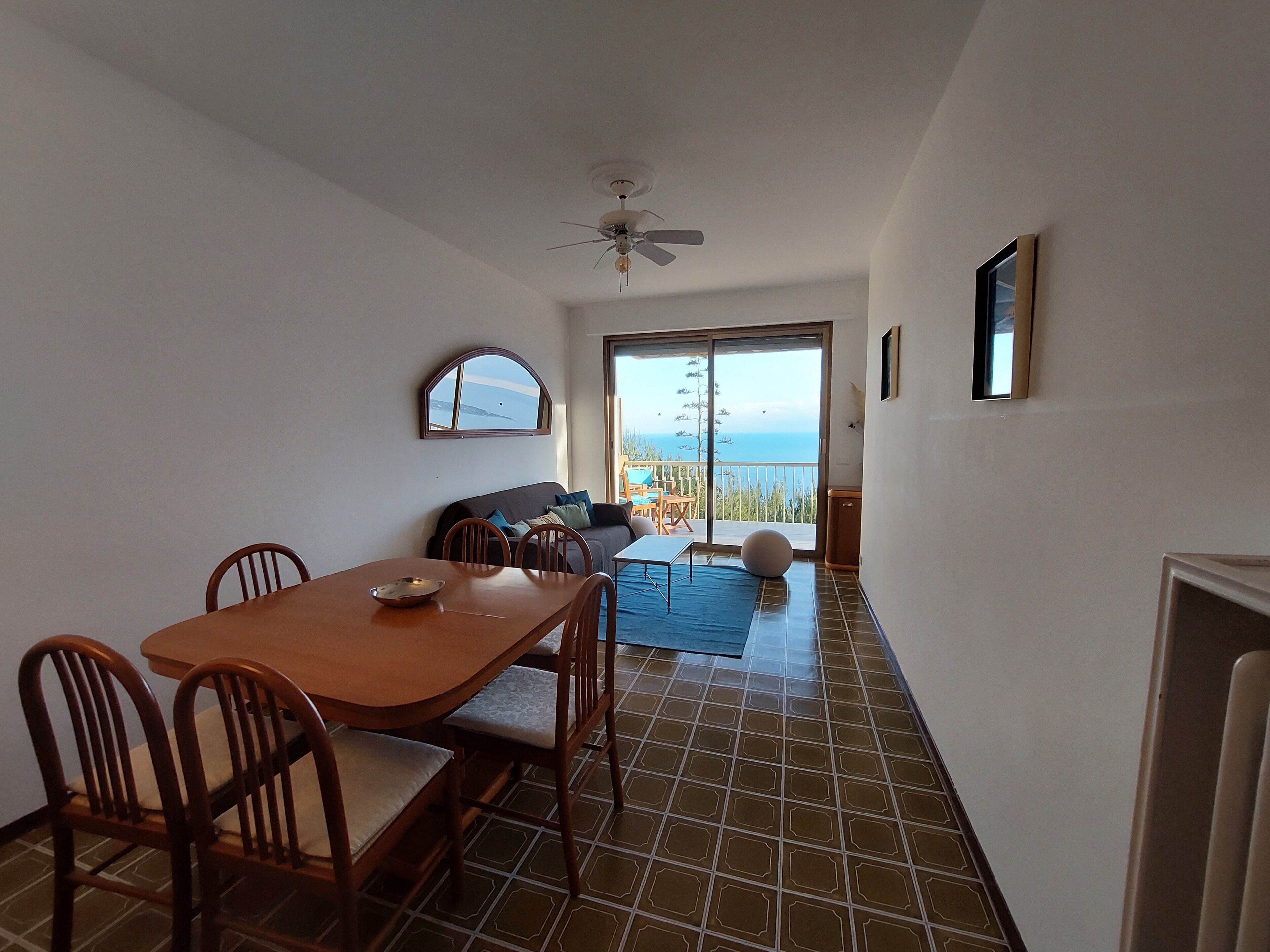 A dining area with a table and chairs, a sofa, and a view of the sea through a sliding door.