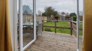 Une vue depuis un balcon doté d’une terrasse en bois, d’une porte vitrée et d’une cour arrière verdoyante, avec une clôture en bois et un abri de jardin.