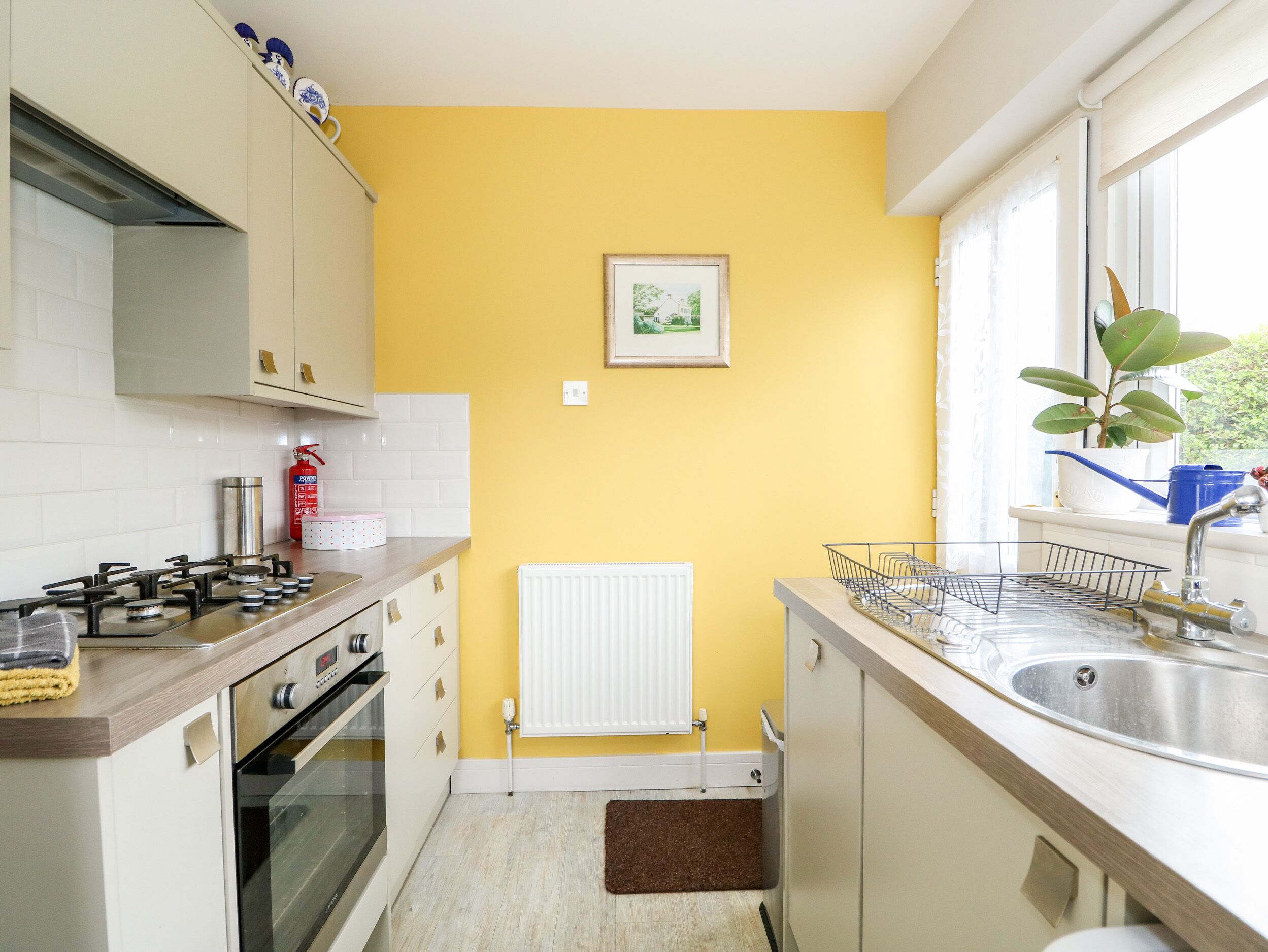 Een compacte keuken met witte kastjes, een gasfornuis, een witte radiator en een raam met een plant.